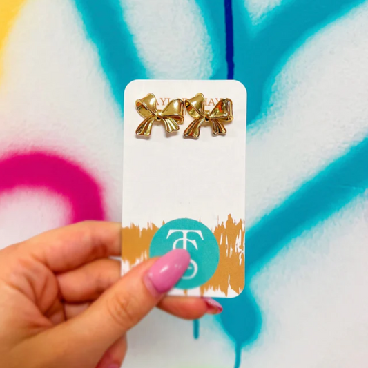 Gold bow-shaped earrings on a card held by a hand against a colorful abstract background