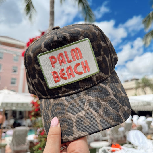 Leopard print cap with 'Palm Beach' text held in front of a tropical setting with palm trees and buildings.