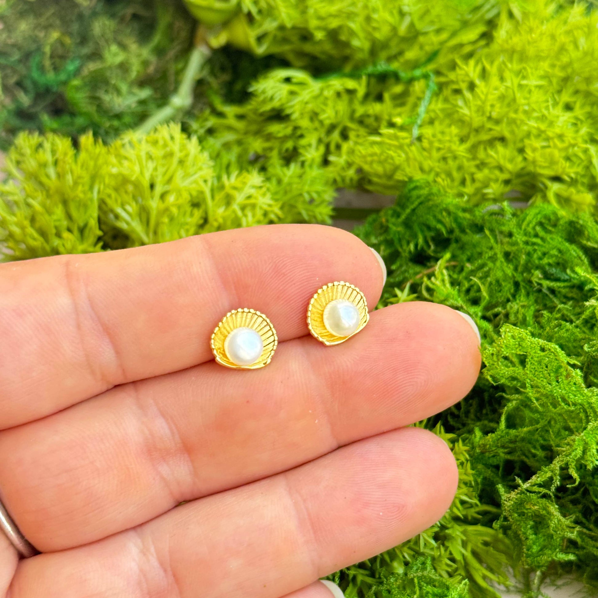 Gold earrings with pearl-like stones held between fingers against a green moss background